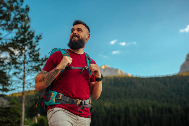 A man with a beard and a red t-shirt is hiking in a mountainous area. He is wearing a backpack and smiling, surrounded by trees and mountains under a clear blue sky.