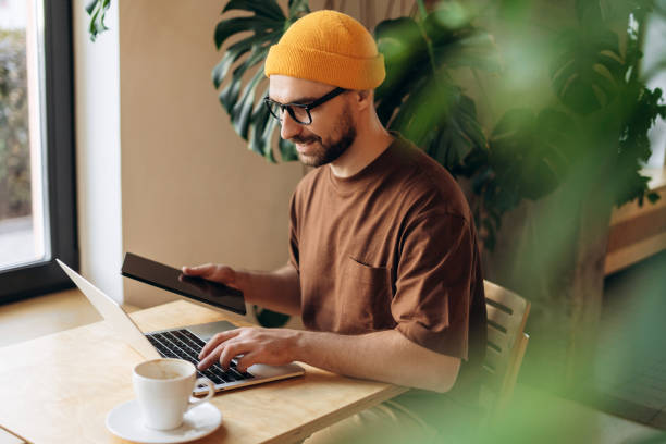istock Happy man, hipster wearing yellow hat and stylish eyeglasses, holding digital tablet, using laptop 2190416393