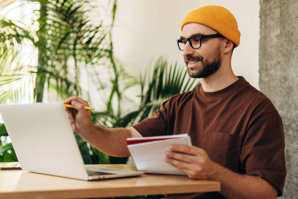 istock Happy successful man, student wearing stylish yellow hat and eyeglasses taking notes, using laptop 2190416416