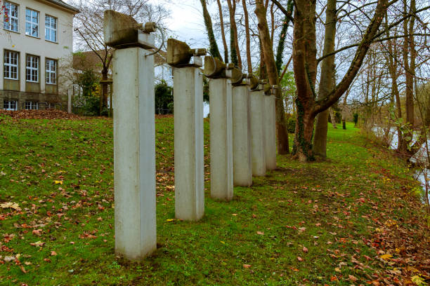 Concrete pillars stand orderly along a grassy area, contrasting against autumn foliage and a tranquil waterway, creating a serene outdoor atmosphere.