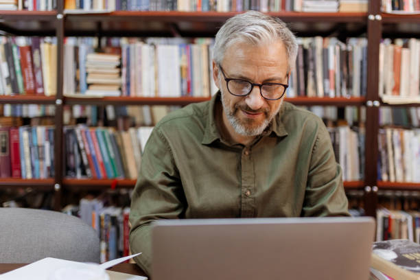 istock Older Man Working on Laptop Surrounded by Books in Library 2190441419