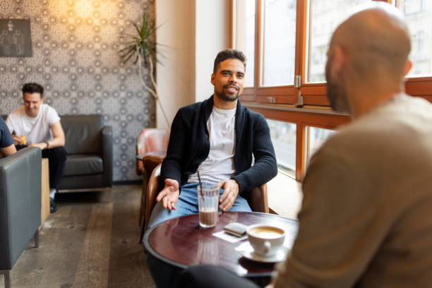 istock Man Sitting by Cafe Window Holding a Drink Talking to his Friend 2190441896