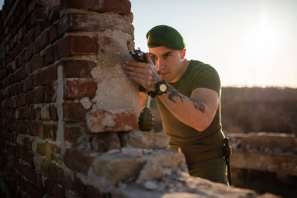 A focused soldier in green beret aims rifle through a broken wall, outdoors during daylight.