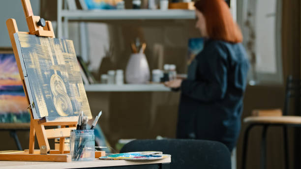 istock Abstract painting black canvas with gold drawings on easel stands on table in workshop studio with paint brushes red-haired unrecognizable woman artist painter choose acrylic oil preparing for drawing 2190461328