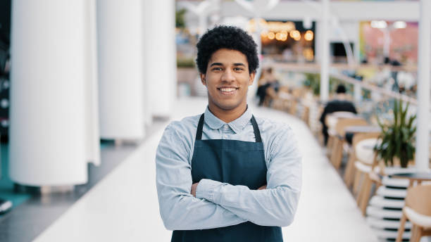 Confident African 30s restaurant worker biracial guy strong position standing indoors barman seller waiter in apron looking to camera successful cafe owner posing with crossed arms small business. High quality 4k footage trabajador de restaurante africano seguro de los años 30, hombre birracial, posición fuerte, de pie en el interior, camarero, vendedor, camarero con delantal, mirando a la cámara, propietario exitoso de un café posando con los brazos cruzados, pequeña
