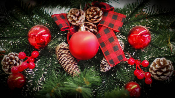 This image beautifully captures a festive holiday wreath featuring a red ornament, plaid ribbon, pinecones, and clusters of red berries set against lush green fir branches. It embodies a cozy and traditional Christmas aesthetic.