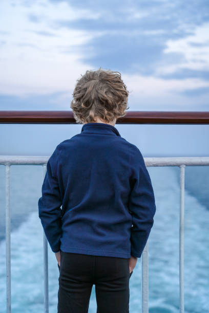 A young child stands at the railing of a cruise ship, looking out over the ocean waves. The soft light of the sunset sky creates a serene atmosphere. Near the coast of Norway