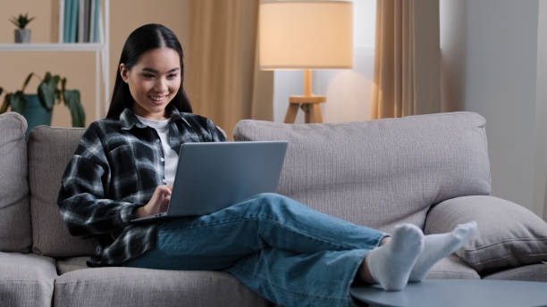 istock Smiling asian woman using laptop casual browsing searching online play computer game. Korean student girl freelancer sitting on couch e-learning internet education chatting studying working with wi-fi 2190478445
