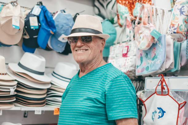A smiling, cheerful senior man tries on a hat while shopping in a store. Elderly witty man looking at camera smiling