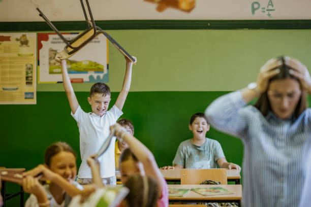 Students are actively engaged in a chaotic moment in class, with excitement and laughter filling the air as one child lifts a chair, drawing attention while others are amused.