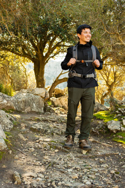 Vertical shot of a cheerful man hiking on a mountain on a rocky path between trees looking around
