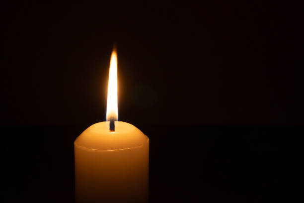 istock A single burning candle flame or light glowing on a big white candle isolated on black or dark background on table in church for Christmas, funeral or memorial service 2190509561