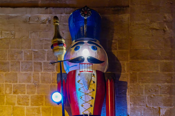 Wooden nutcracker soldier with bright red and yellow attire, illuminated by colorful blue and warm lights, stands against textured stone wall