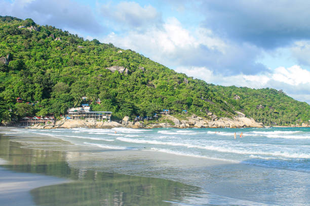 istock Famous Haad Rin Beach in Koh Phangan with white sandy shoreline, rolling waves, and beachfront resorts nestled against green hillside with granite boulders 2190510376