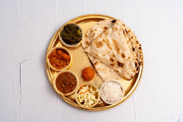 A beautifully presented Indian thali served on a golden plate. This meal includes freshly made naan bread, steamed basmati rice, creamy lentil curry (dal), mixed vegetable curry, paneer masala, and saag (spinach dish). A small portion of coleslaw adds freshness, while a round, bright orange gulab jamun offers a sweet finish to this vibrant and balanced meal. The image showcases the richness and variety of Indian cuisine in an elegant and festive presentation