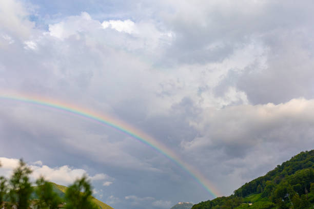 A vibrant rainbow stretches across the sky above rolling green hills under grey clouds.