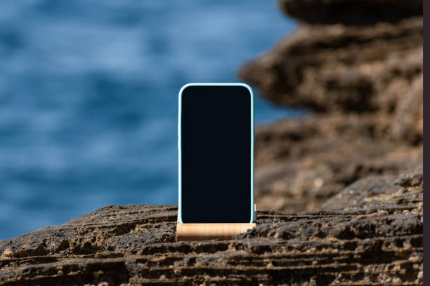 Mobile phone with black blank screen mockup on rocky sea coast, blue water background, copy space. Phone device mock-up. Cellular network Internet coverage, mobile signal, remote travel destination