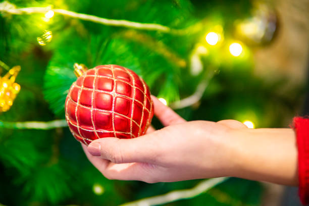 Close up of woman's hand holding a shiny red Christmas ball hanging on decorated Xmas tree.