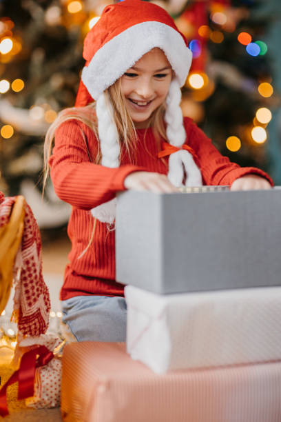 istock Young girl unwrapping Christmas gift with joyful expression near Christmas tree 2190523959