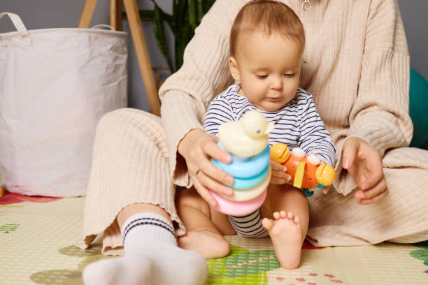 istock A small child sits on a soft mat, focused on stacking colorful rings and holding a toy, while a caregiver engages nearby in a warm home setting 2190524921