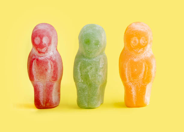 Three gummy Jelly Babies  stand upright in red, green, and orange colors against a bright yellow background. Each has a sugary, frosted coating and playful expressions.