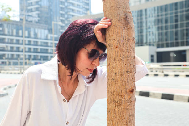 A woman leaning against a tree on a noisy street, overcome with heartache, seeks help from passersby, thinking about her health