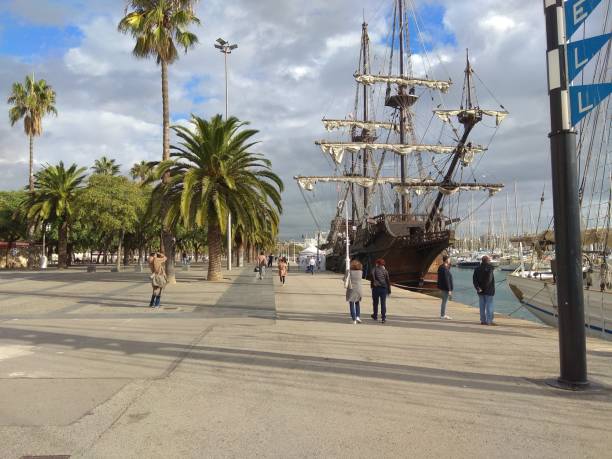 Barcelona, ​​Spain - 08.11.2018: A picturesque view of a historic sailing ship docked at a lively marina, surrounded by palm trees and a bustling promenade. The ship's tall masts and intricate rigging stand against a dramatic sky, while people stroll along the waterfront enjoying the scenic beauty. This vibrant scene captures a blend of maritime history and modern leisure, creating a perfect destination for visitors and photographers