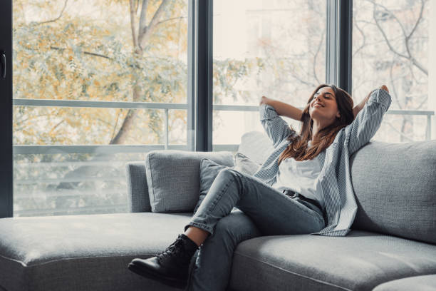 Relaxed serene pretty young woman feel fatigue lounge on comfortable sofa hands behind head rest at home, happy calm lady dream enjoy wellbeing breathing fresh air in cozy home modern living room