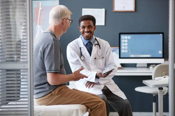 Smiling Black Doctor Holding Tablet Talking to Senior Patient in Consultation Smiling Black Doctor Holding Tablet Talking to Senior Patient in Consultation