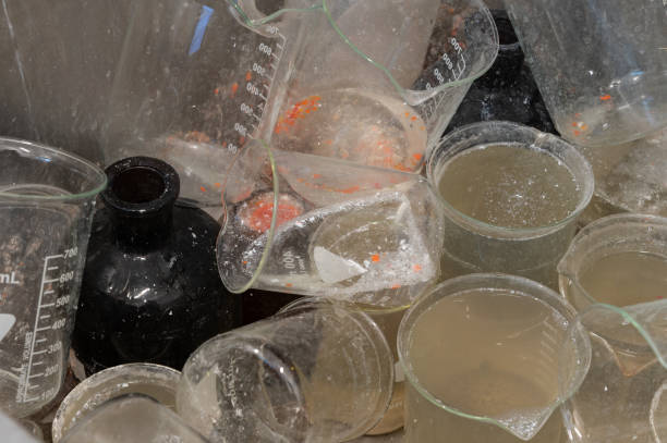 A collection of used laboratory glassware, including beakers and flasks, covered in residue and containing murky liquids. The scene reflects post-experiment cleanup in a scientific or industrial setting.