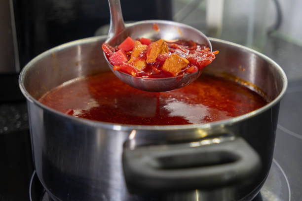 Ladle scooping hot borscht with vegetables from a large pot on a kitchen stove