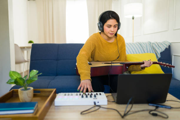 Young latin woman playing guitar and using a midi keyboard while composing music at home using a laptop