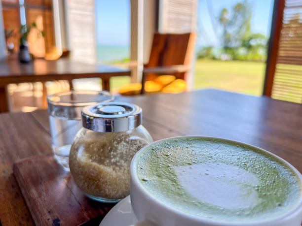Matcha latte and sugar on a wooden table with ocean view. Spacious cafe with open windows and tropical setting.
