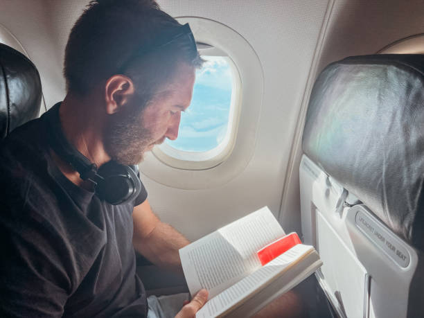 istock Man Reading a Book on Airplane Near Window 2190620442