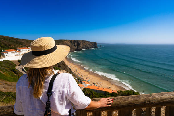 istock Mature woman tourist in summer hat standing at viewpoint overlooking beach between cliffs - Arrifana, Algarve, Portugal. Back view 2190631130