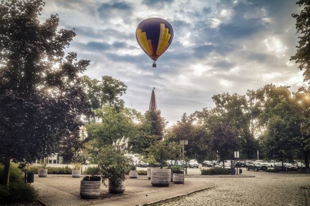 A vibrant hot air balloon floats over a serene urban park with lush greenery and a cloudy sky.