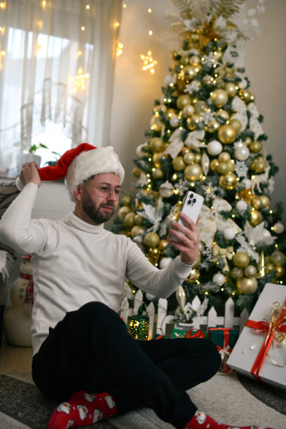 istock Man Sitting on Ground With Crossed Legs Under Christmas Tree Capturing Selfie With Smartphone 2190664520