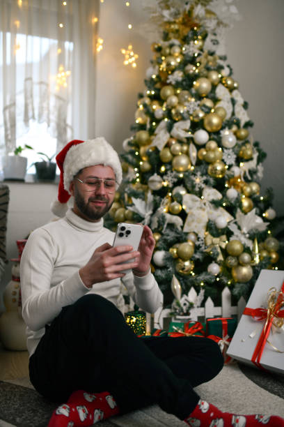 istock Handsome Male Sitting Under Christmas Tree Wearing Santa Hat and Text Messaging 2190664578