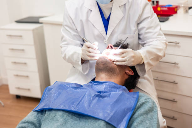 Dentist wearing a mask and gloves is performing a dental check-up on a male patient lying on a dental chair in a brightly lit clinic, using specialized tools and a focused light