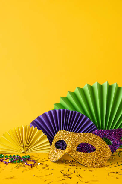 istock Mardi Gras side vertical view themed setup showcasing a glittery golden mask, colorful beads, and paper fans against a vibrant yellow background 2190668191