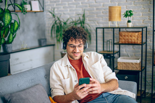 istock Young happy man with headphones using his smartphone while relaxing in the couch at home 2190671420