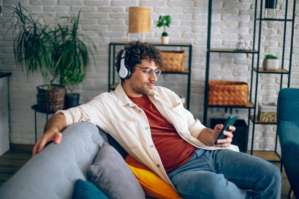 istock Young happy man with headphones using his smartphone while relaxing in the couch at home 2190671431