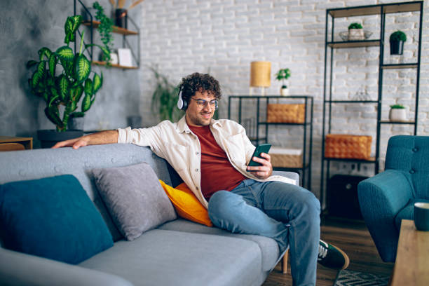 istock Young happy man with headphones using his smartphone while relaxing in the couch at home 2190671447