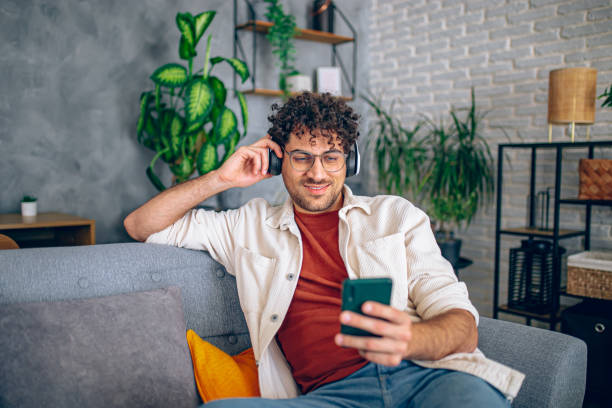 istock Young happy man with headphones using his smartphone while relaxing in the couch at home 2190671468