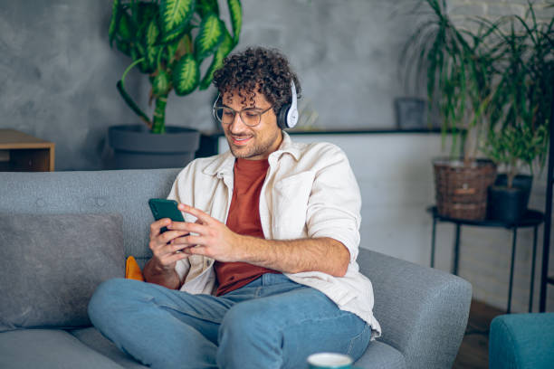 istock Young happy man with headphones using his smartphone while relaxing in the couch at home 2190671734