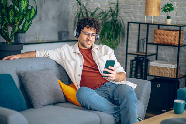 istock Young happy man with headphones using his smartphone while relaxing in the couch at home 2190671766
