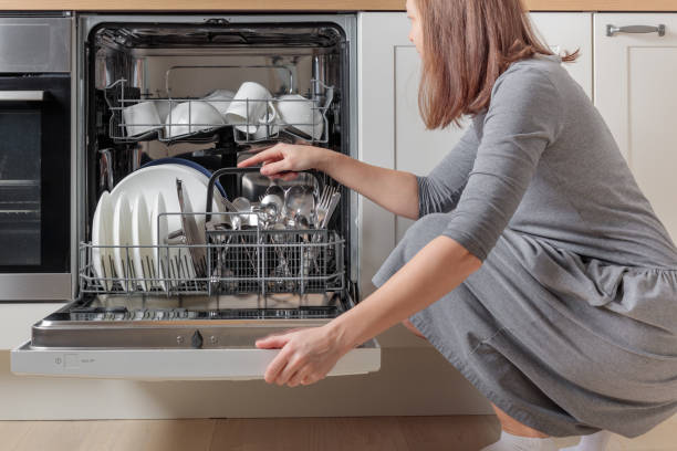 istock Woman's hands emptying, unloading a dishwasher, taking out a cutlery basket with spoons, knives and forks. Household chores with a modern kitchen appliance. An open dishwasher with white plates and stainless steel cutlery 2190674462