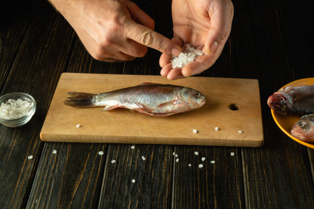 istock Skilled hands sprinkle coarse salt over a fresh fish on a wooden cutting board, setting the stage for a flavorful dish. Nearby, additional fish await their turn in the kitchen 2190677051