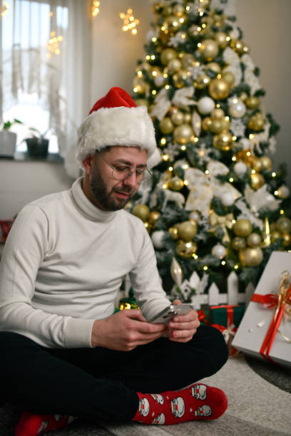 istock Handsome Male With Santa Hat Sitting in Front of Christmas Tree Checking Messages on Mobile Device 2190683243