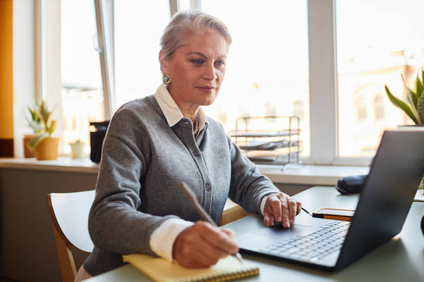 istock Senior Female Employee Using Laptop while Writing in Notepad Indoors 2190684200